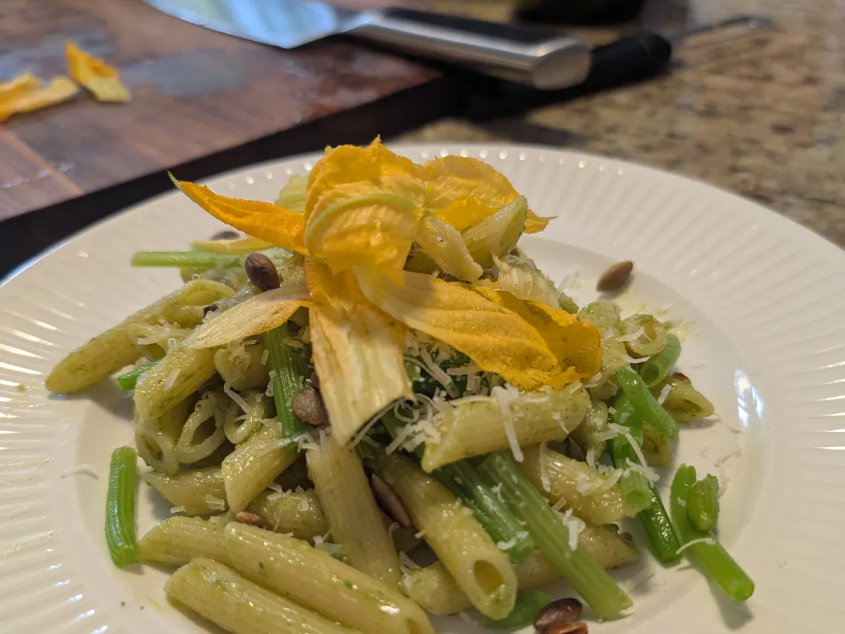 Assiette de penne aux tiges de zucchini garnie de pétales de fleurs de zucchini, de pecorino et de graines grillées, avec une planche et un couteau en arrière-plan