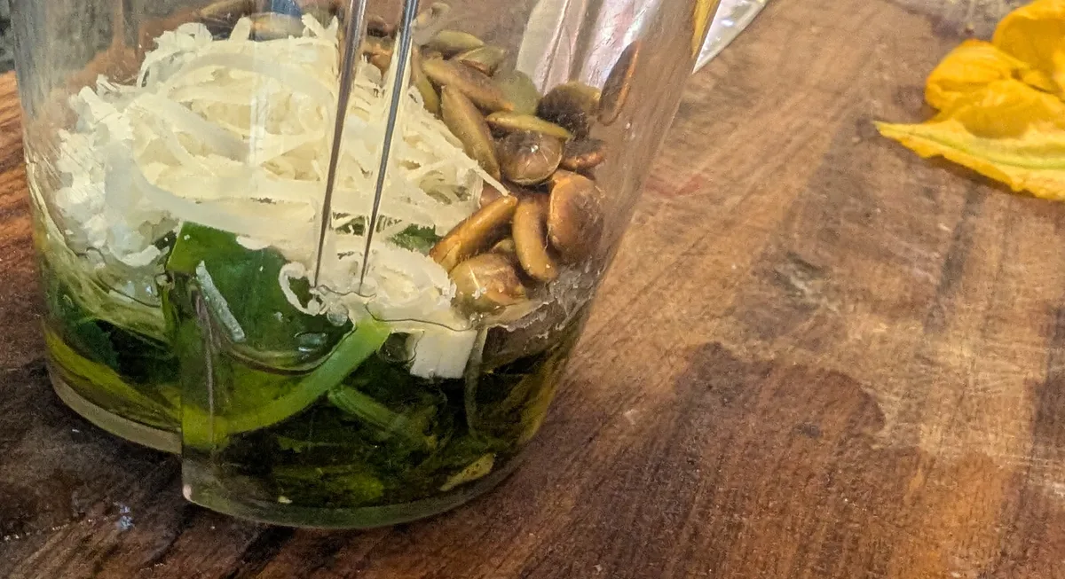 Récipient du mélangeur contenant des feuilles de zucchini blanchies, de l'huile, du pecorino râpé et des graines de citrouille grillées, avec une fleur de zucchini sur la planche