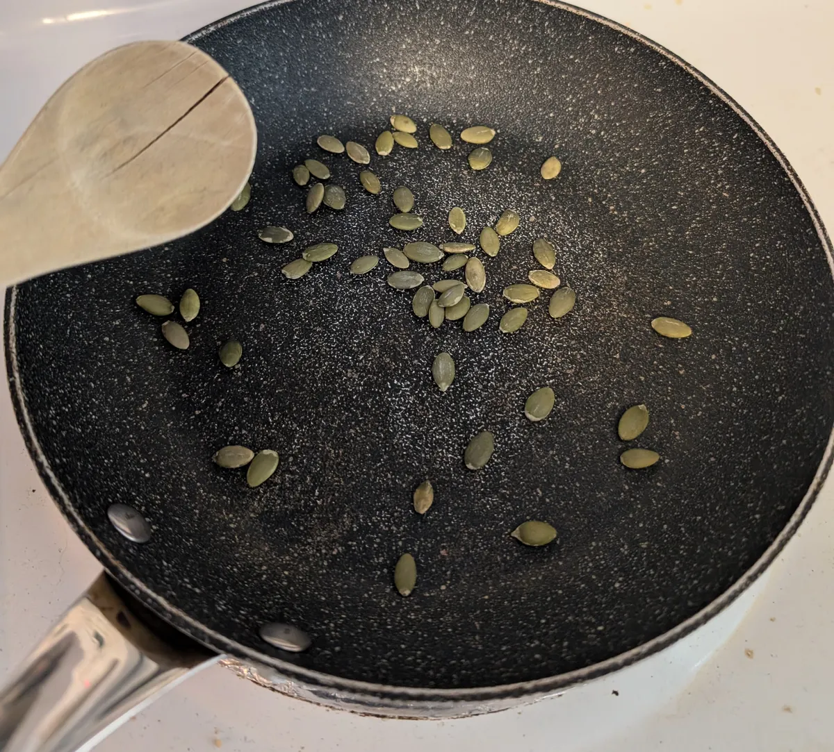 Graines de citrouille vertes en train de griller à sec dans une poêle antiadhésive, avec une cuillère de bois